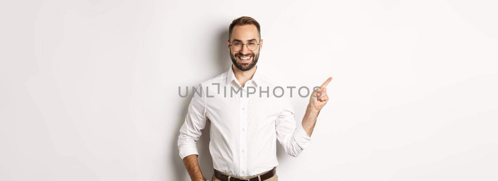Confident businessman pointing finger left at company logo, standing over white background.