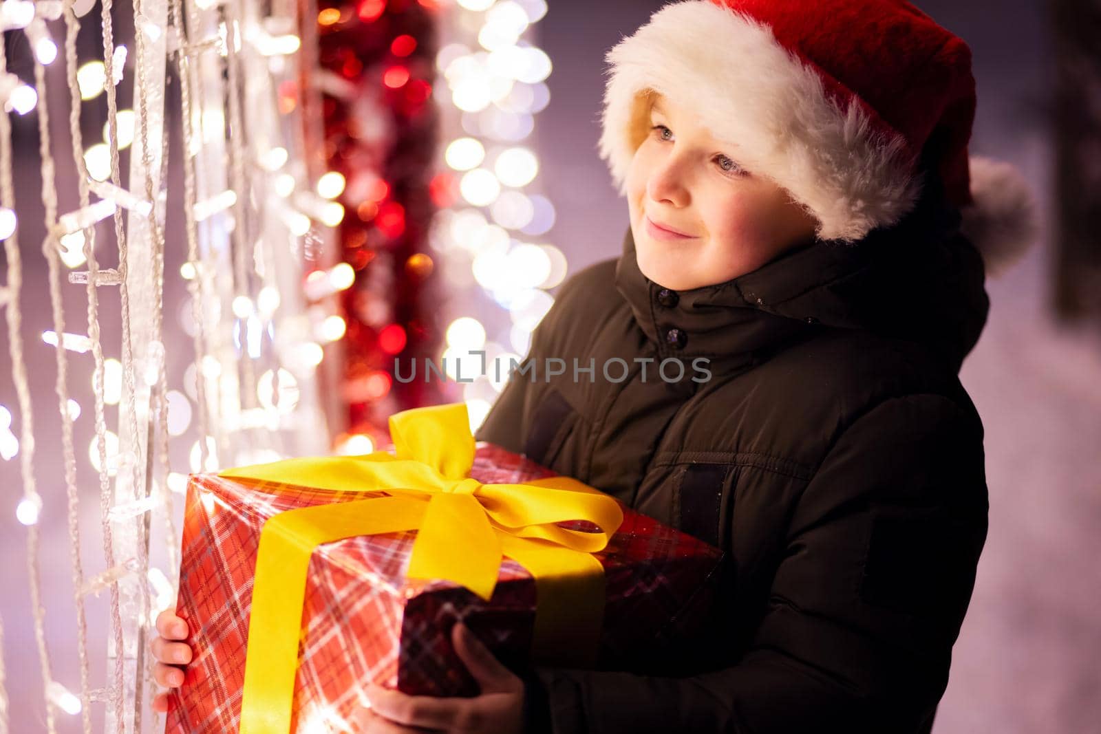 Happy child in a red Santa Claus hat with wrapped Christmas gift near shining light bulbs in winter evening. The concept of Christmas and New Year.