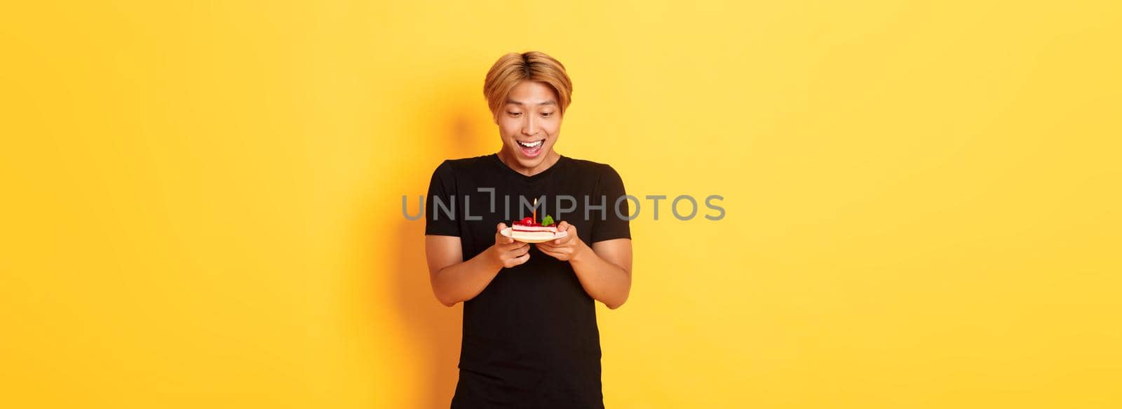 Excited happy attractive asian guy smiling as looking at birthday cake, making wish, celebrating b-day, standing yellow background.