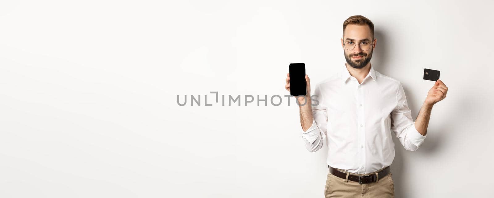 Handsome bearded man showing mobile phone and credit card, shopping online, standing over white background.