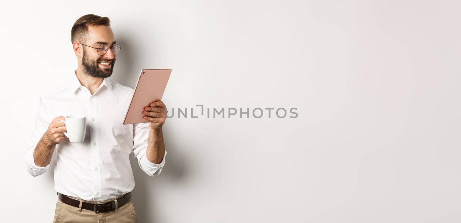 Handsome businessman drinking coffee and reading on digital tablet, smiling pleased, standing over white background.