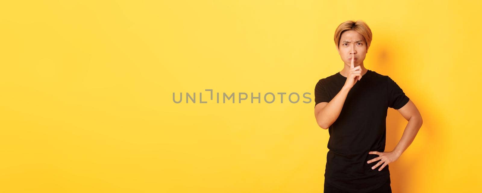 Portrait of angry asian man frowning, shushing at camera disappointed, standing yellow background by Benzoix