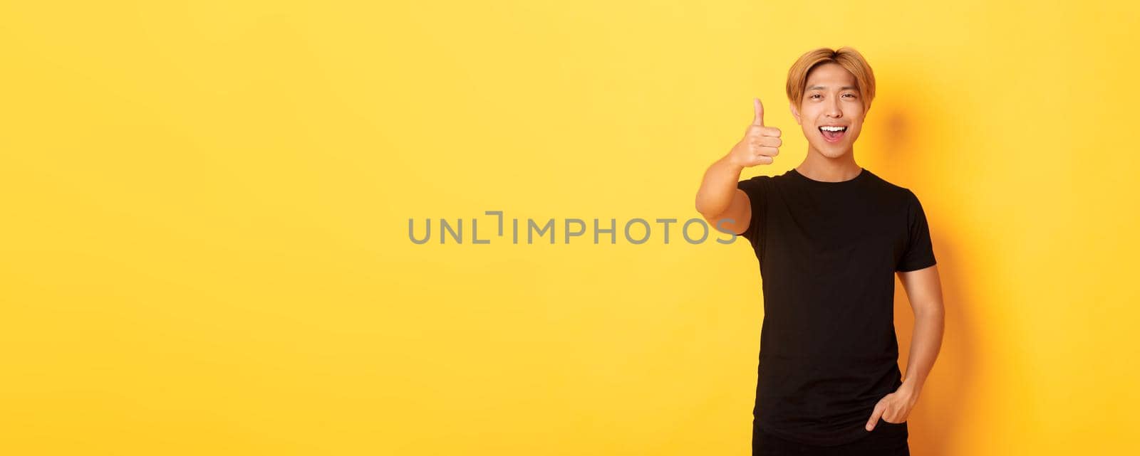 Portrait of pleased handsome asian guy, showing thumbs-up in approval, standing over yellow background.