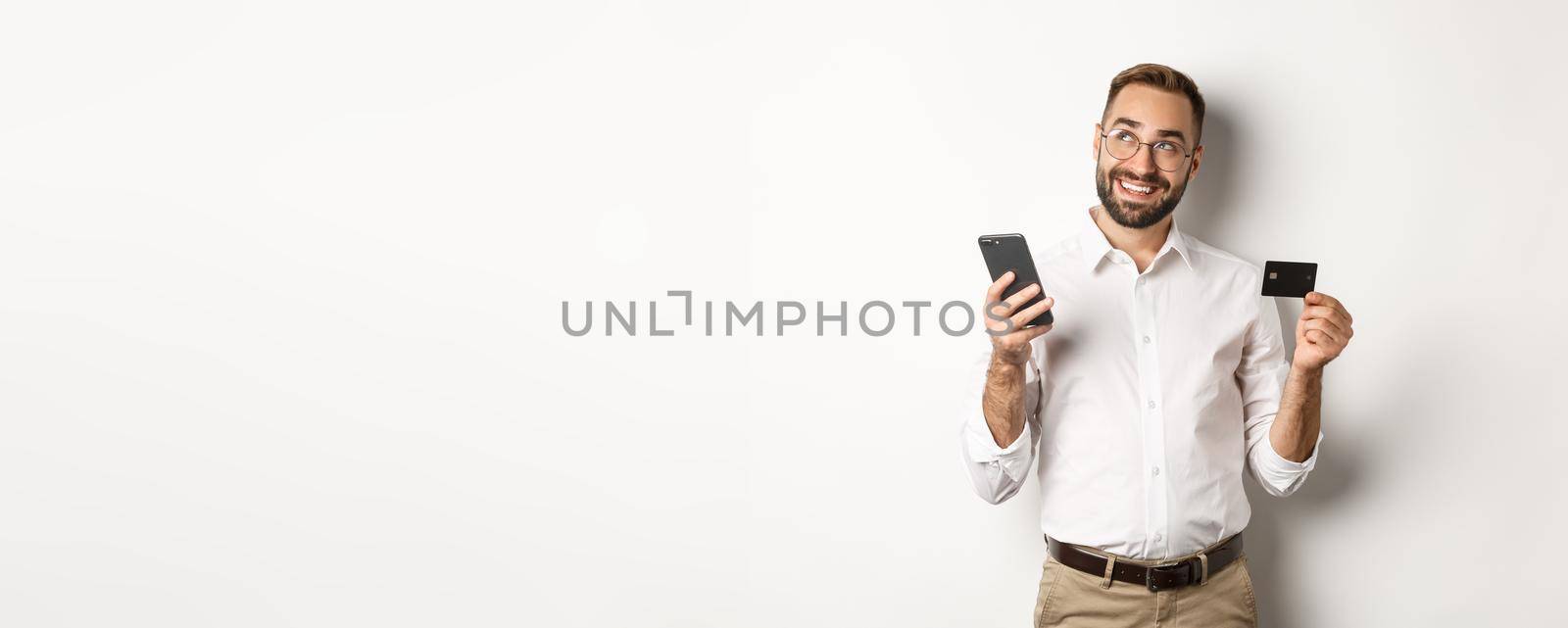 Business and online payment. Image of handsome man thinking while holding credit card and smartphone, standing against white background.