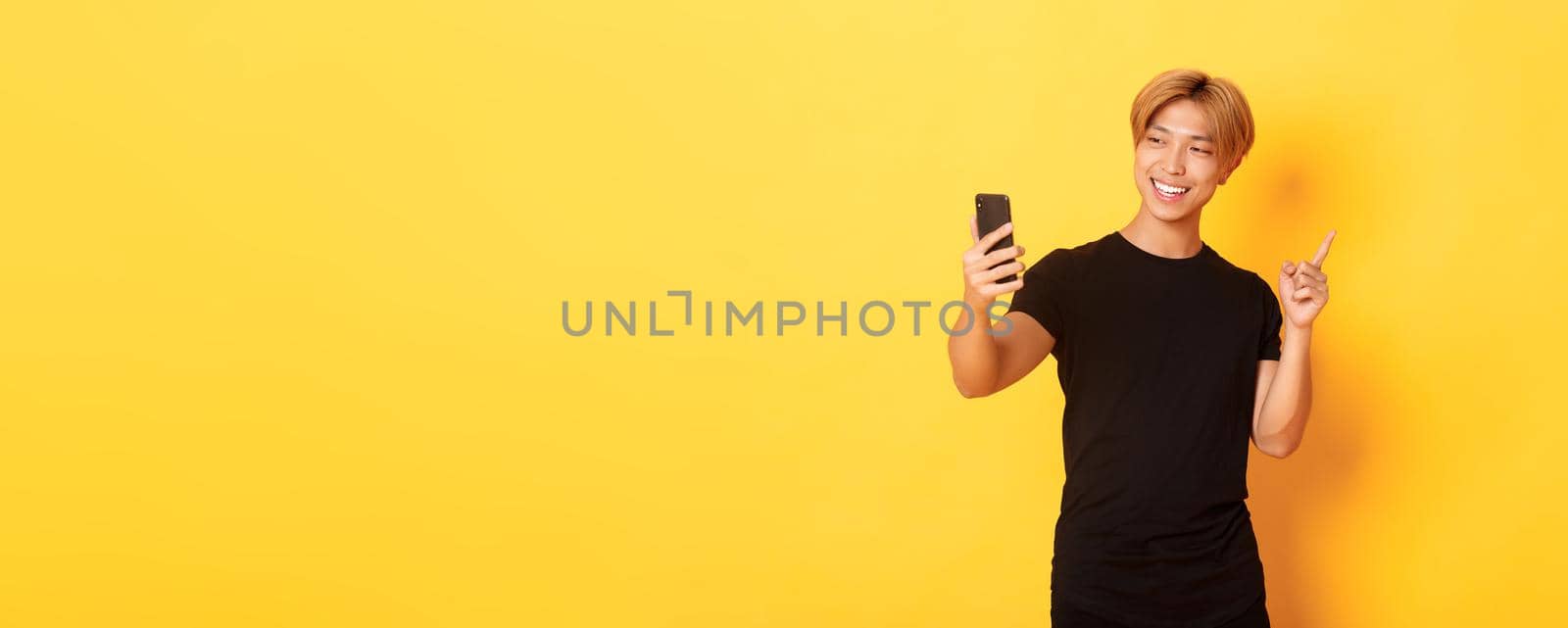 Portrait of handsome smiling asian man having video call on smartphone and pointing finger at something, standing yellow background by Benzoix