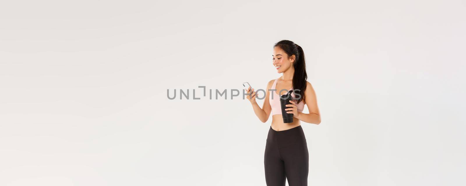 Full length of smiling fit and healthy female fitness coach, asian sportswoman checking water tracker reminder in smartphone, using application during workout in gym, white background by Benzoix