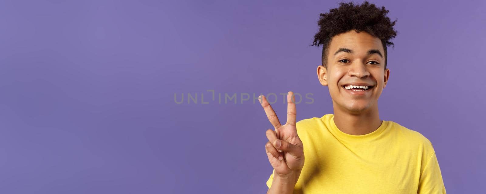 Close-up portrait of handsome upbeat young teenage guy with afro hairstyle, show peace sign and smiling, wear yellow t-shirt, staying optimistic and positive, purple background.