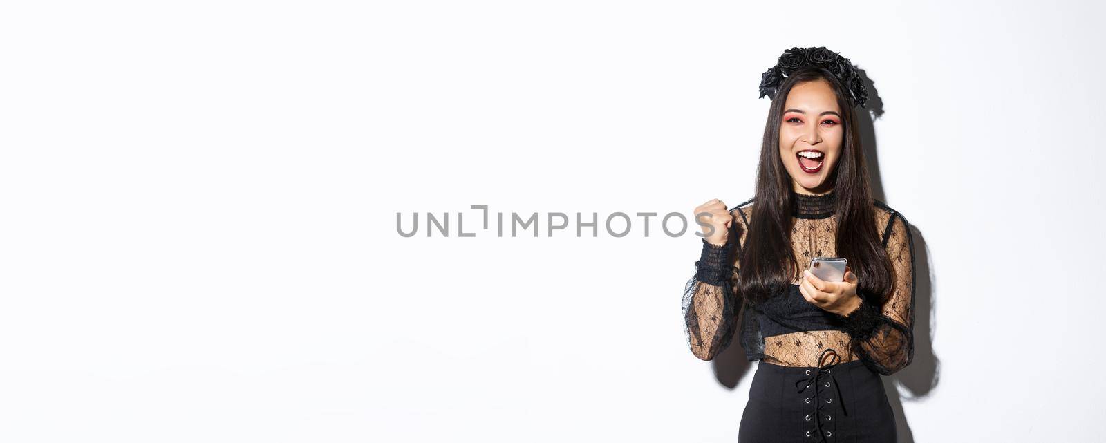 Image of successful happy asian woman celebrating good news, fist pump in rejoice and holding smartphone, wearing halloween costume, standing over white background by Benzoix