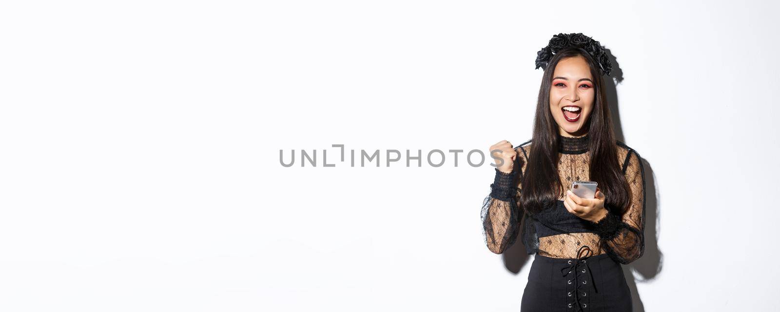 Image of successful happy asian woman celebrating good news, fist pump in rejoice and holding smartphone, wearing halloween costume, standing over white background.