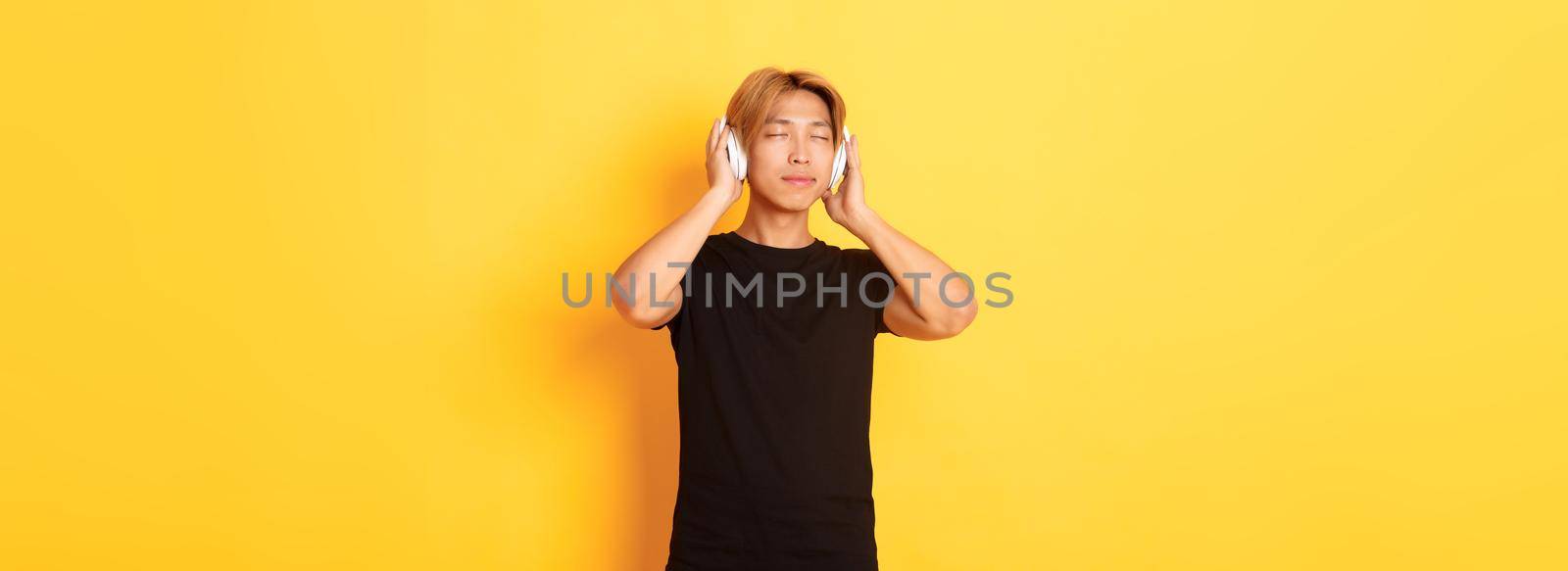 Relaxed and pleased handsome asian guy close eyes from satisfaction while listening music in wireless headphones, standing yellow background.