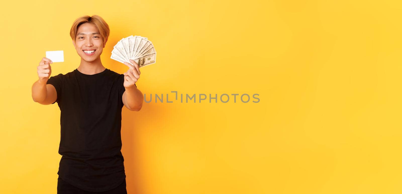 Satisfied attractive korean guy with blond hair, showing money and credit card, smiling happy, yellow background.