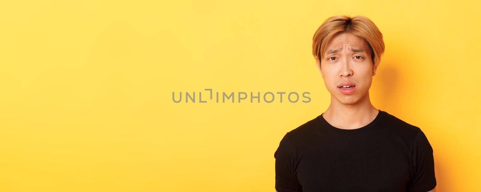Close-up of confused and upset asian blond guy looking puzzled, open mouth and frowning perplexed, standing over yellow background.