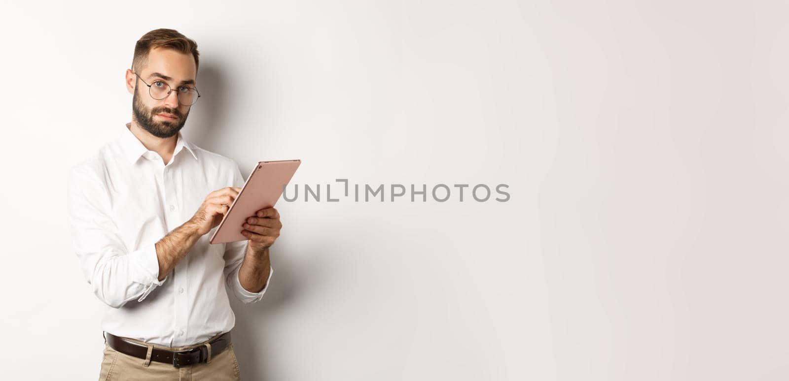 Handsome businessman doing job on digital tablet, reading something, standing over white background.
