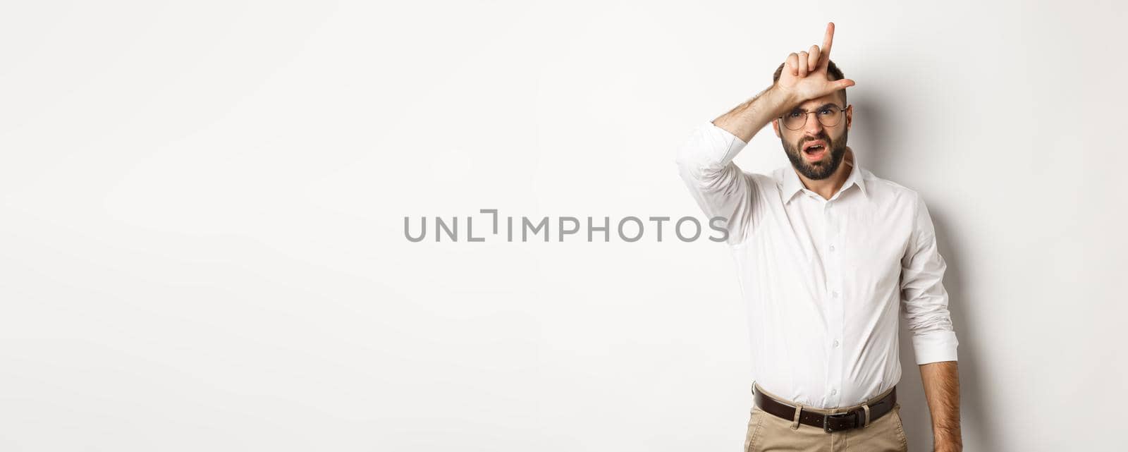 Shocked guy showing loser sign on forehead, complaining, standing over white background.