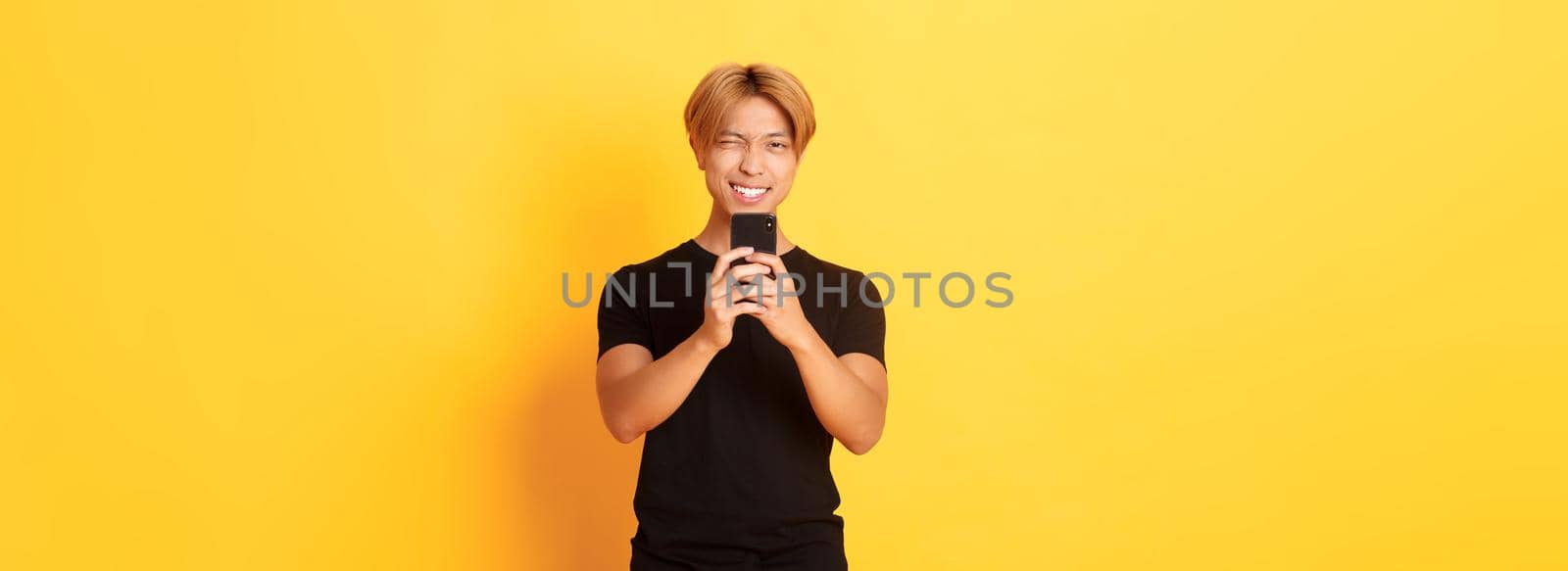 Stylish handsome asian guy taking picture on smartphone and smiling, photographing with mobile phone, standing yellow background.