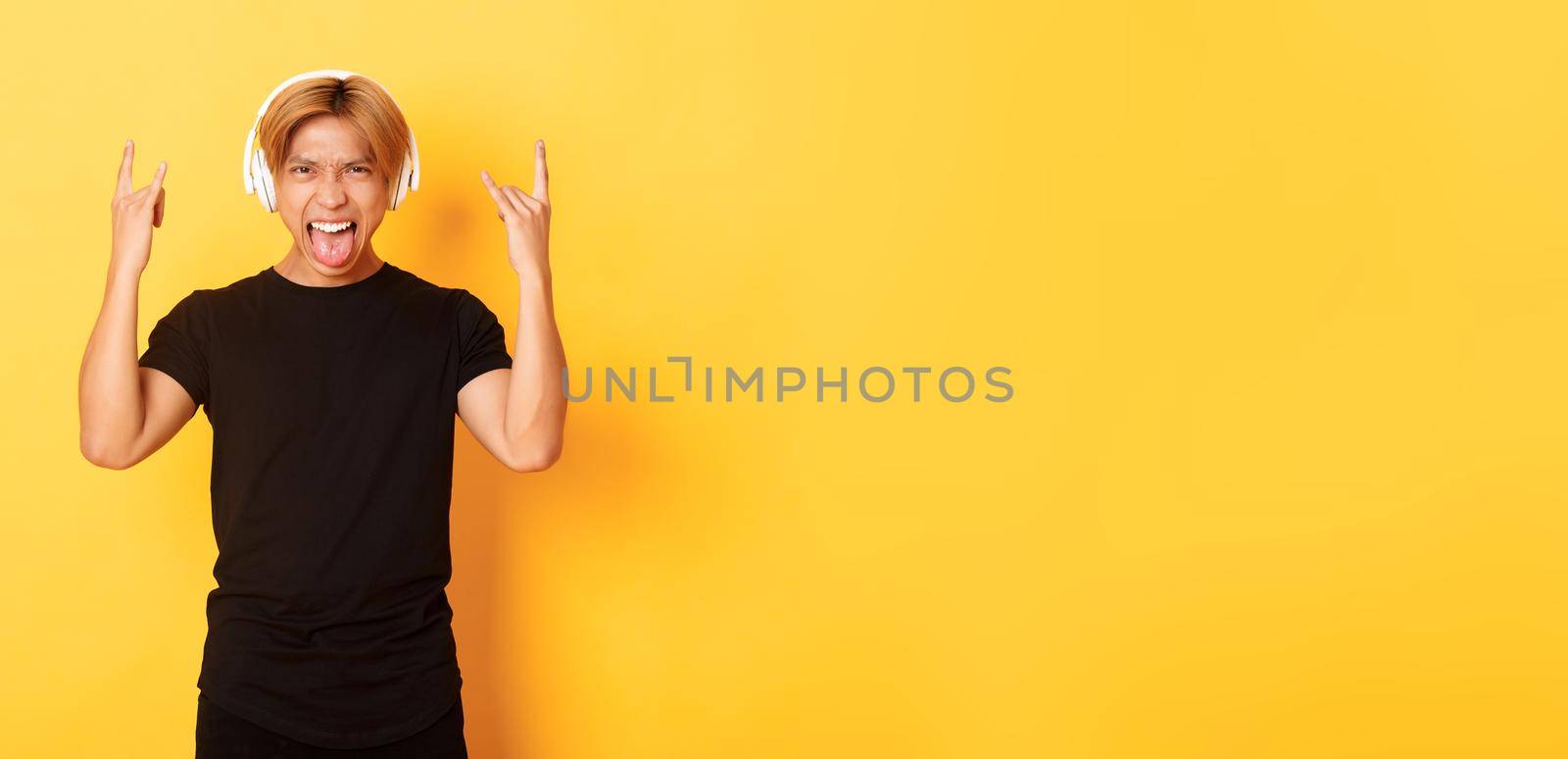 Sassy handsome asian guy likes heavy metal, listening hard rock music, showing rock-n-roll gesture and wearing wireless headphones, sticking tongue, standing yellow background by Benzoix