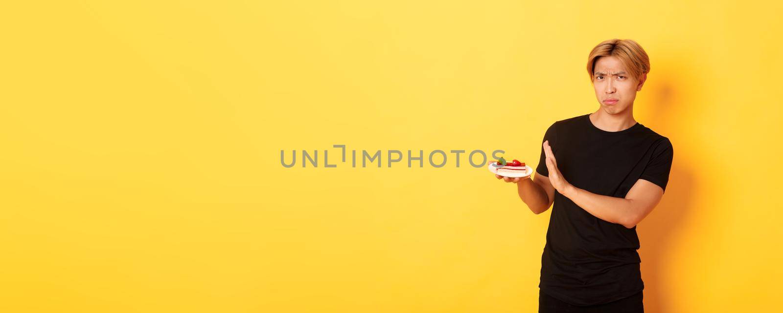 Portrait of disgusted and upset young asian guy refuse eating cake, looking displeased and showing rejection gesture, yellow background.