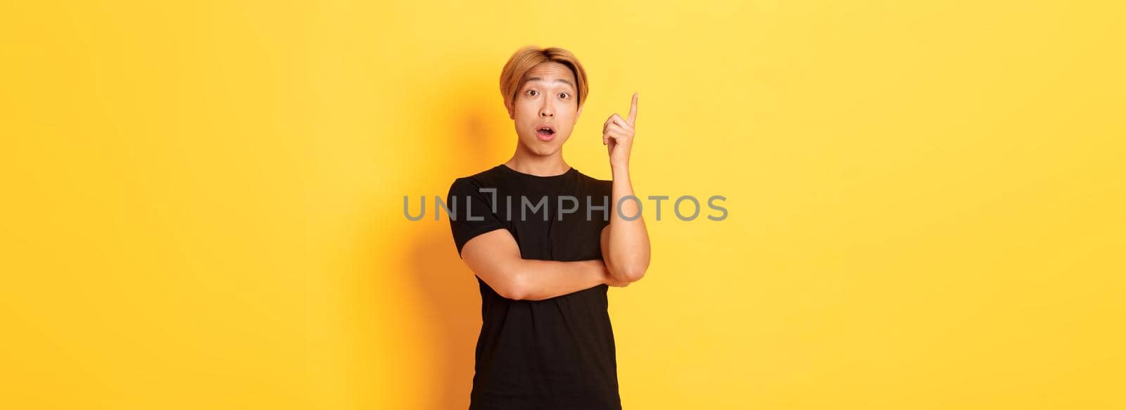 Portrait of thoughtful handsome asian man raising finger, suggesting idea, standing yellow background by Benzoix