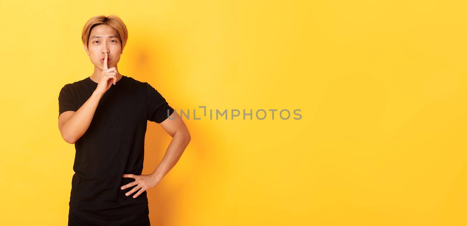 Portrait of attractive blond asian man shushing, press finger to lips and hush, yellow background by Benzoix
