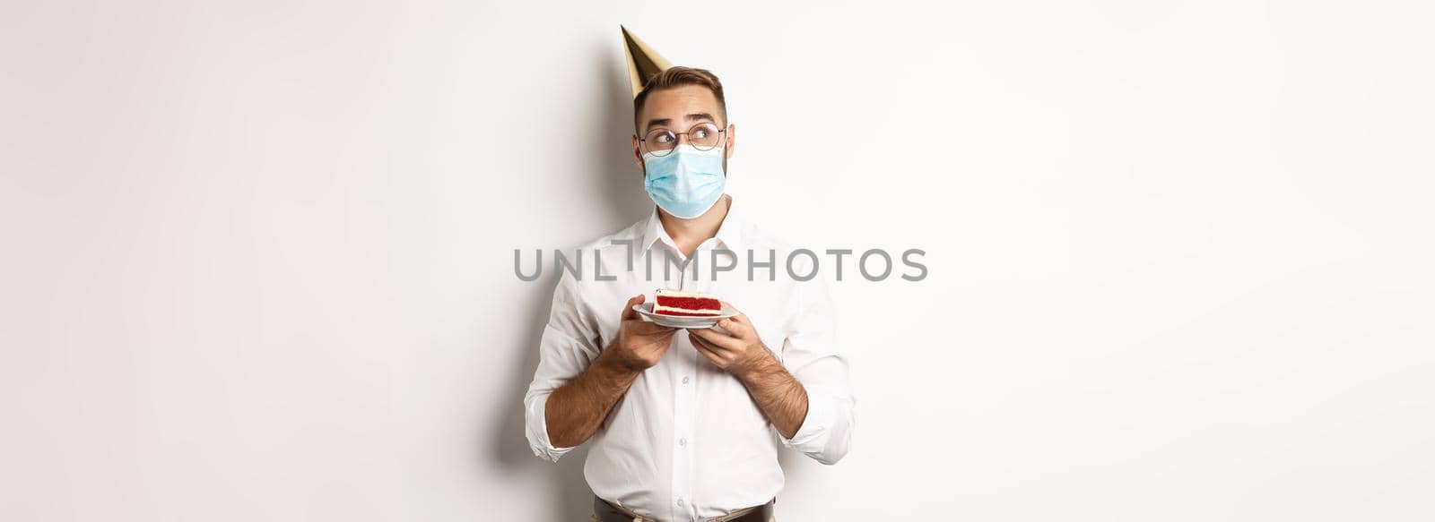 Covid-19, social distancing and celebration. Thoughtful man holding birthday cake, making wish and wearing face mask on quarantine, white background.