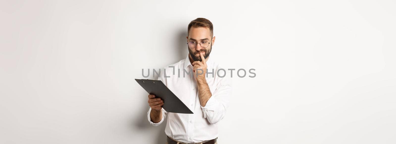 Thoughtful employee looking at clipboard, having doubts, standing over white background by Benzoix