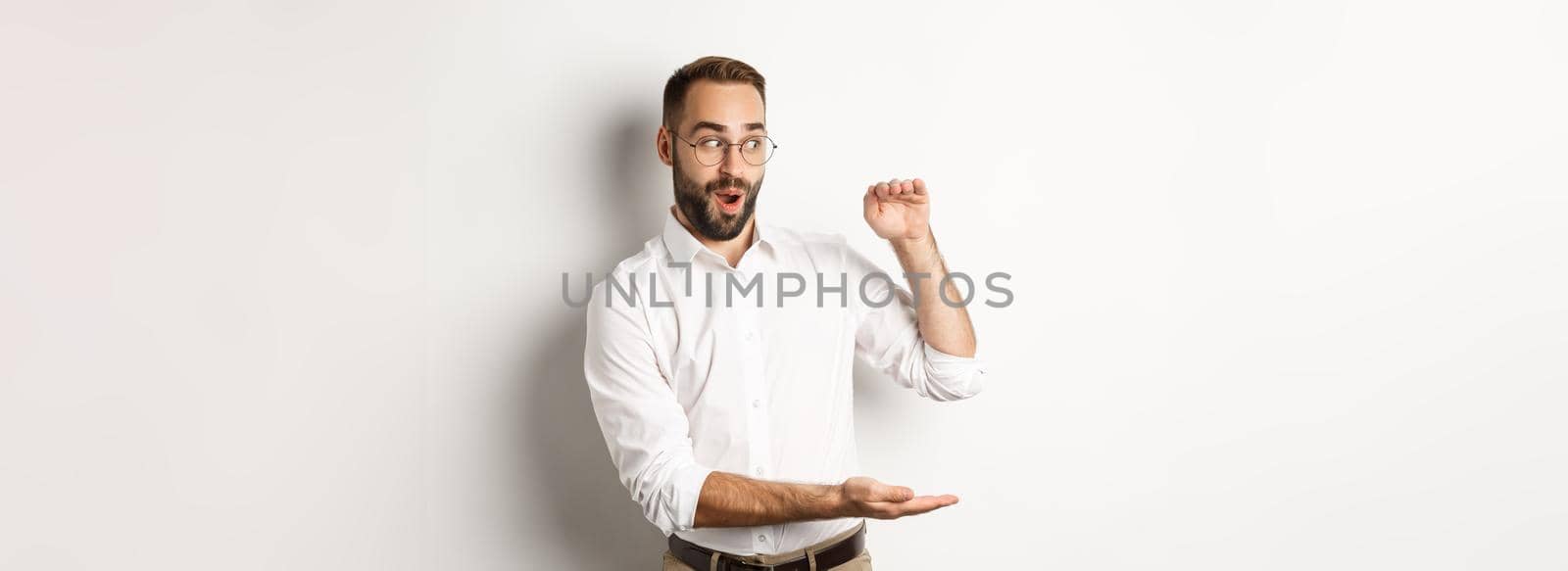 Amazed businessman showing big object, describe something large and looking excited, standing over white background.