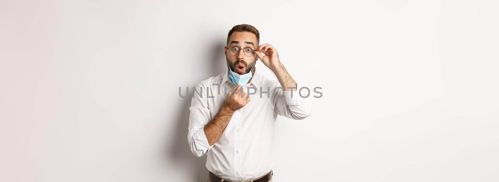 Covid-19, social distancing and quarantine concept. Impressed business man take off medical mask, looking surprised, standing over white background.