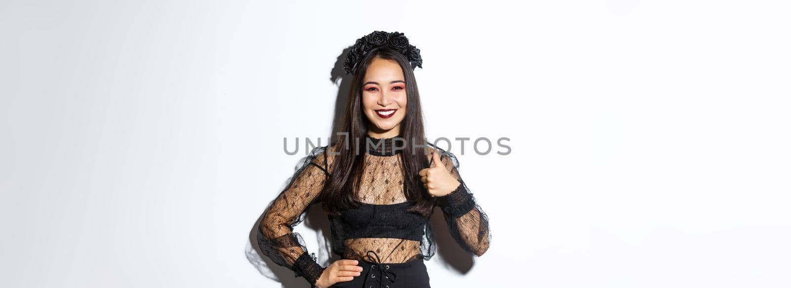 Image of satisfied pretty asian girl in halloween costume showing thumbs-up in approval, smiling pleased. Woman in evil witch party dress looking happy, like something, white background.