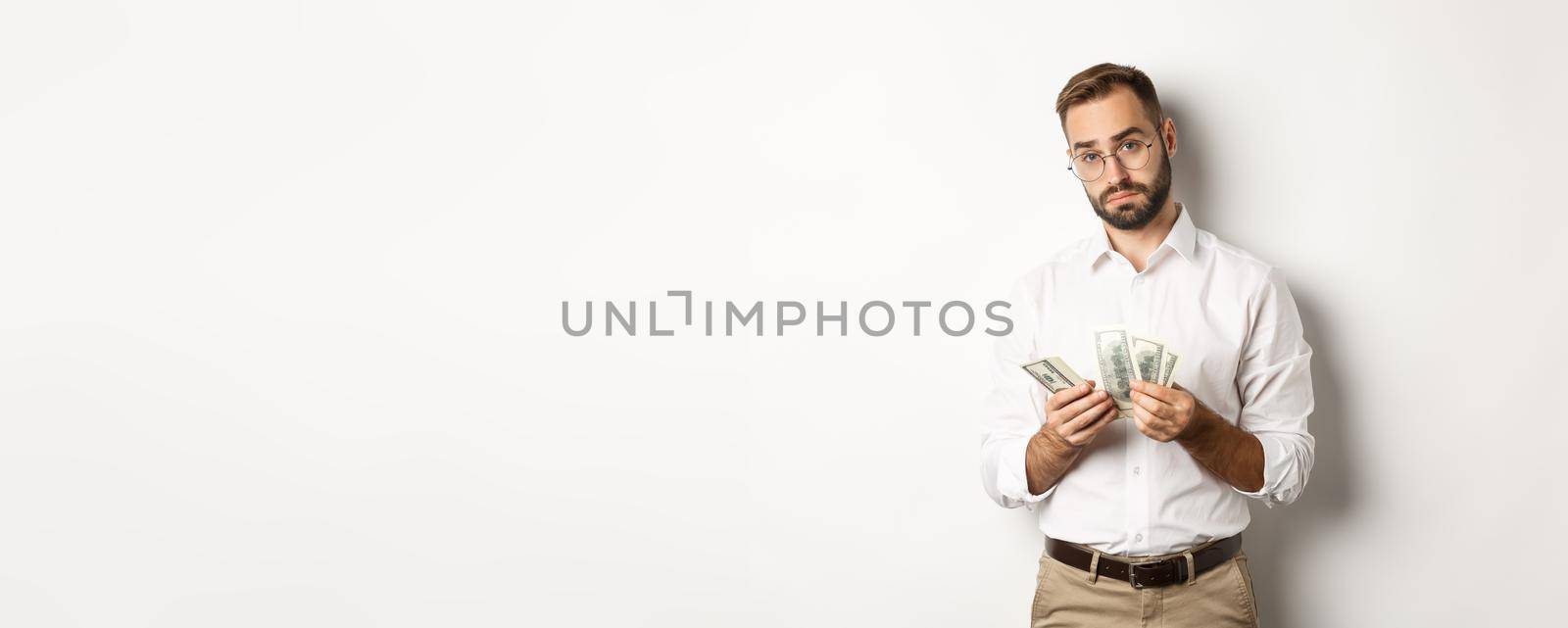 Handsome businessman counting money and looking at camera, standing serious against white background.