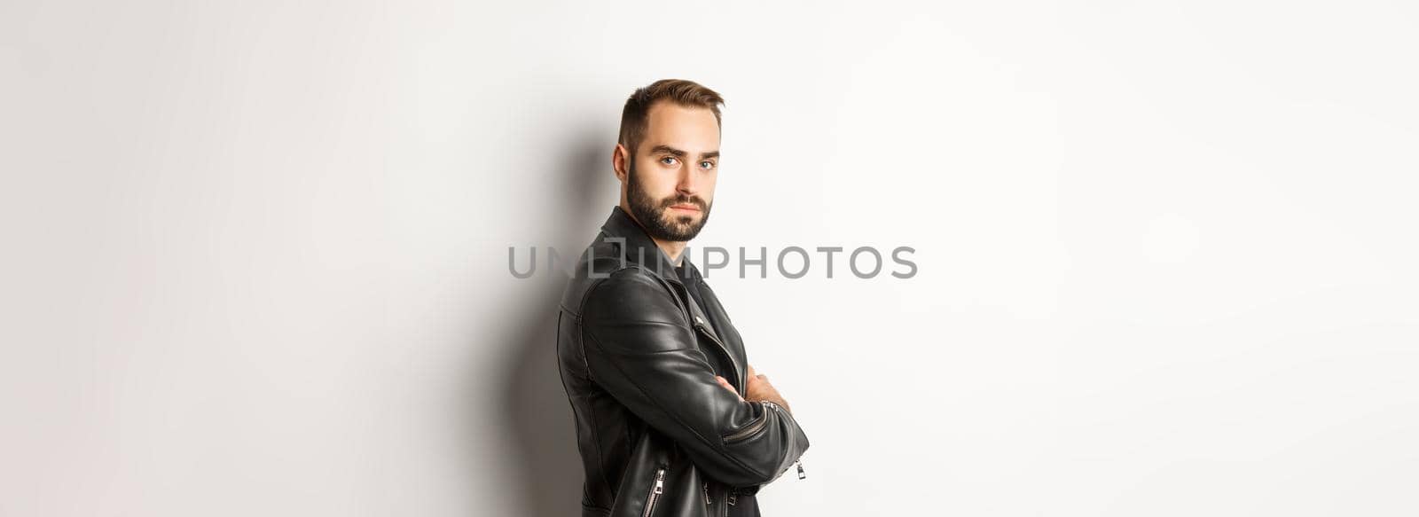 Profile of confident and handsome bearded man in black leather jacket, turn face at camera and looking serious, white background.