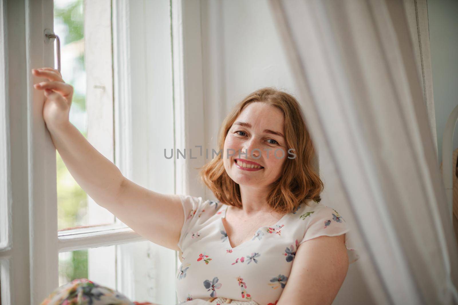 A middle-aged woman in a cream dress sits mysteriously and looks out the window on the windowsill. Green trees outside. by Matiunina