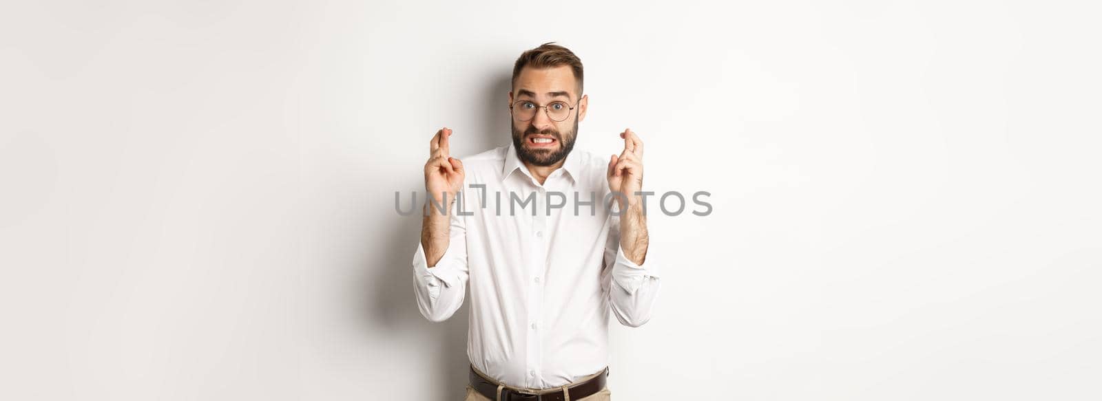 Worried man making a wish, cross fingers and hope for relish, standing over white background.