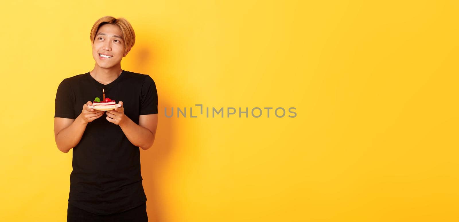 Portrait of handsome dreamy asian guy looking upper left corner and thinking, making wish while celebrating birthday and holding b-day cake, yellow background.