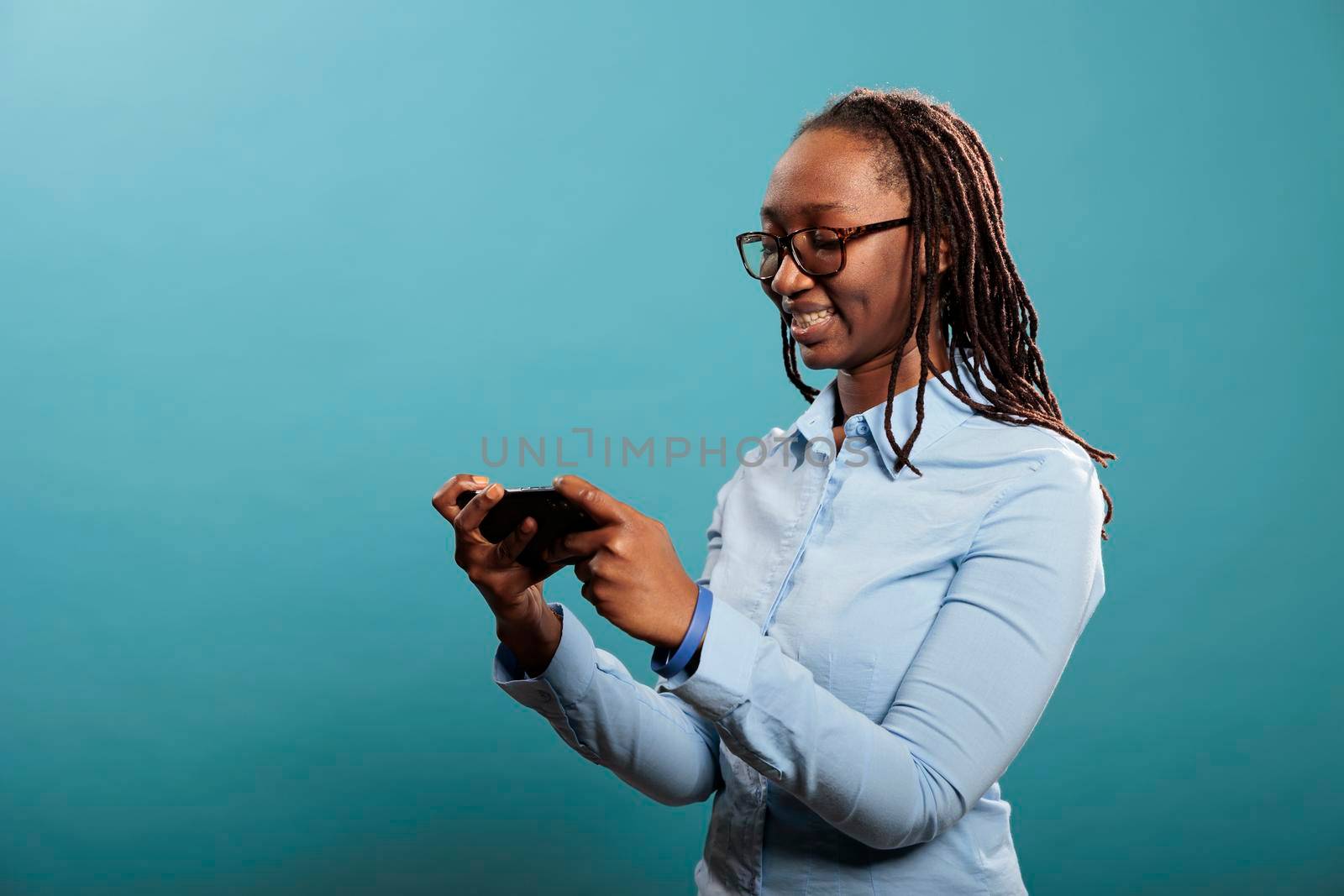 Beautiful happy woman with touchscreen smartphone browsing internet webpages while standing on blue background. Confident joyful young adult person with modern cellphone watching video content.