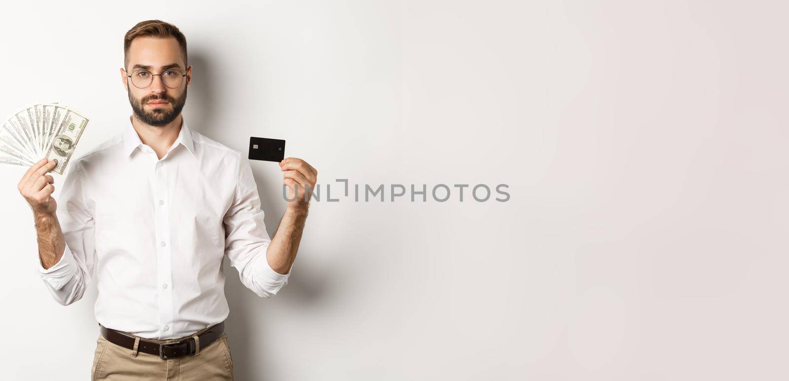 Serious businessman looking at camera, holding credit card and money, standing over white background. Concept of shopping and finance by Benzoix