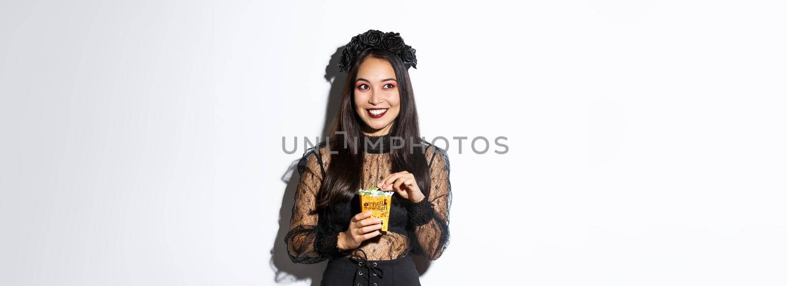 Smiling cute asian woman celebrating halloween, holding sweets and grinning happy, trick or treating in witch costume.