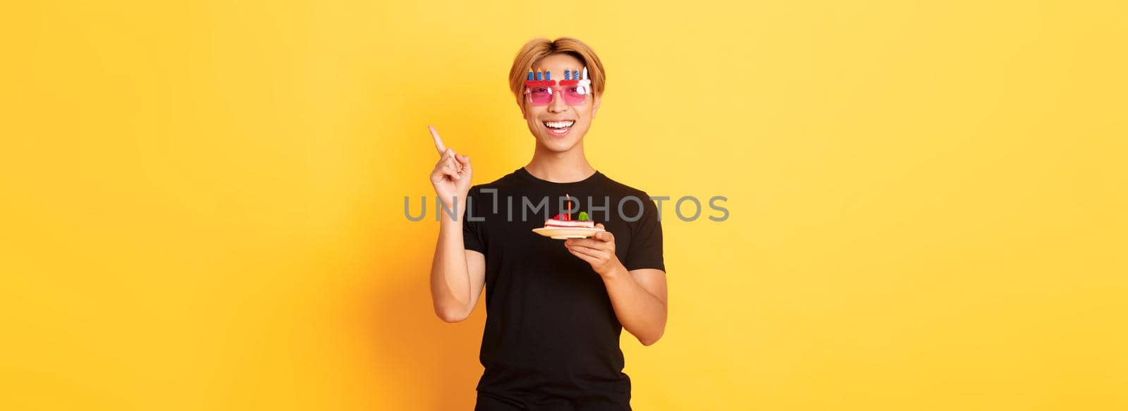 Cheerful, handsome asian birthday guy in funny sunglasses, holding b-day cake with candle and pointing finger upper right corner at your logo, yellow background.
