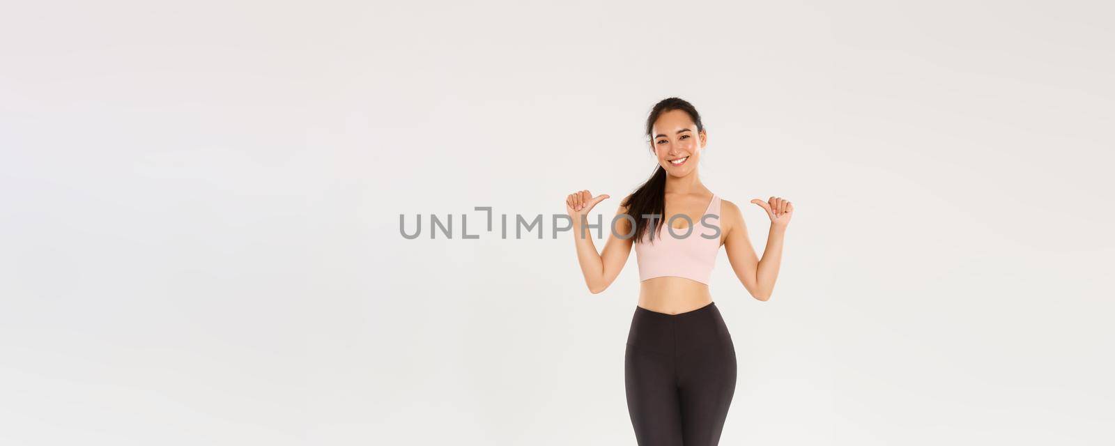 Full length of professional fitness coach, asian girl athelte pointing at herself and smiling happy, workout in gym, gaining perfect body with training, wearing sportswear, white background.