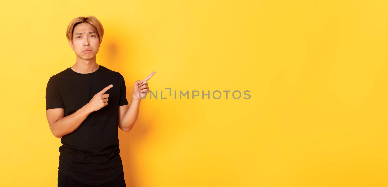 Portrait of disappointed and sad handsome blond asian guy, sulking upset, pointing fingers upper right corner, standing yellow background.