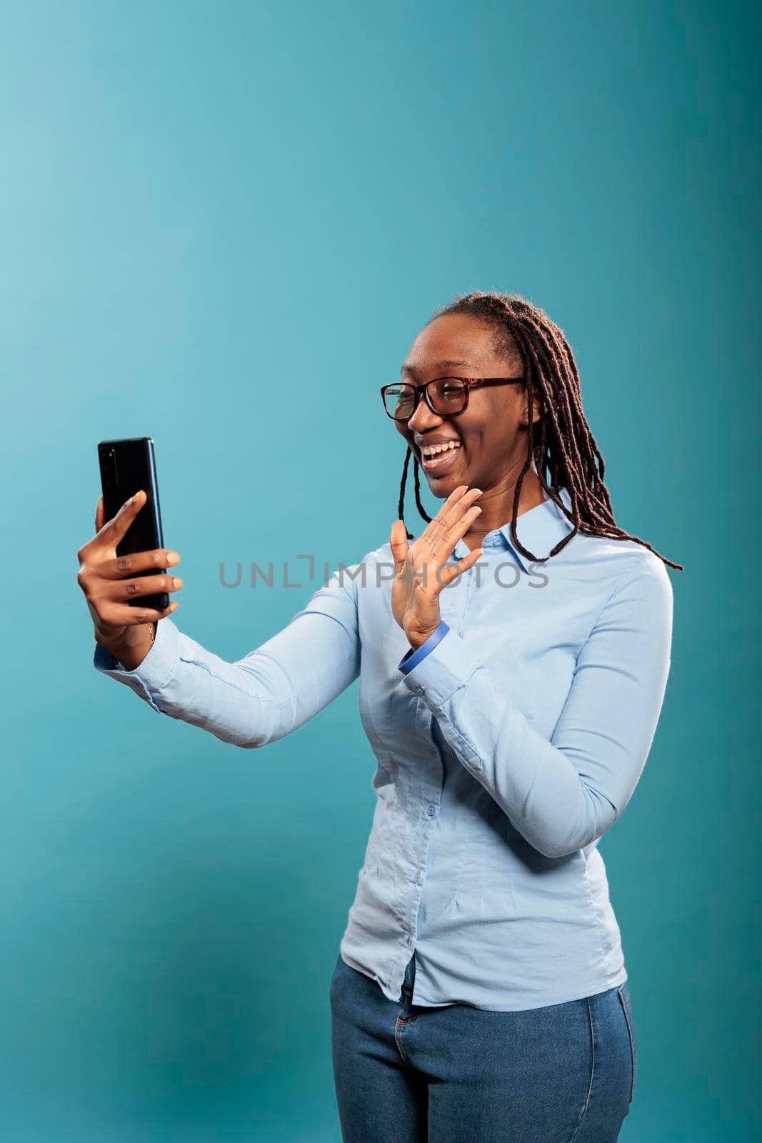 Beautiful confident young lady greeting friend over online phone video call while standing on blue background. Happy smiling african american woman with smartphone waving at video conference call