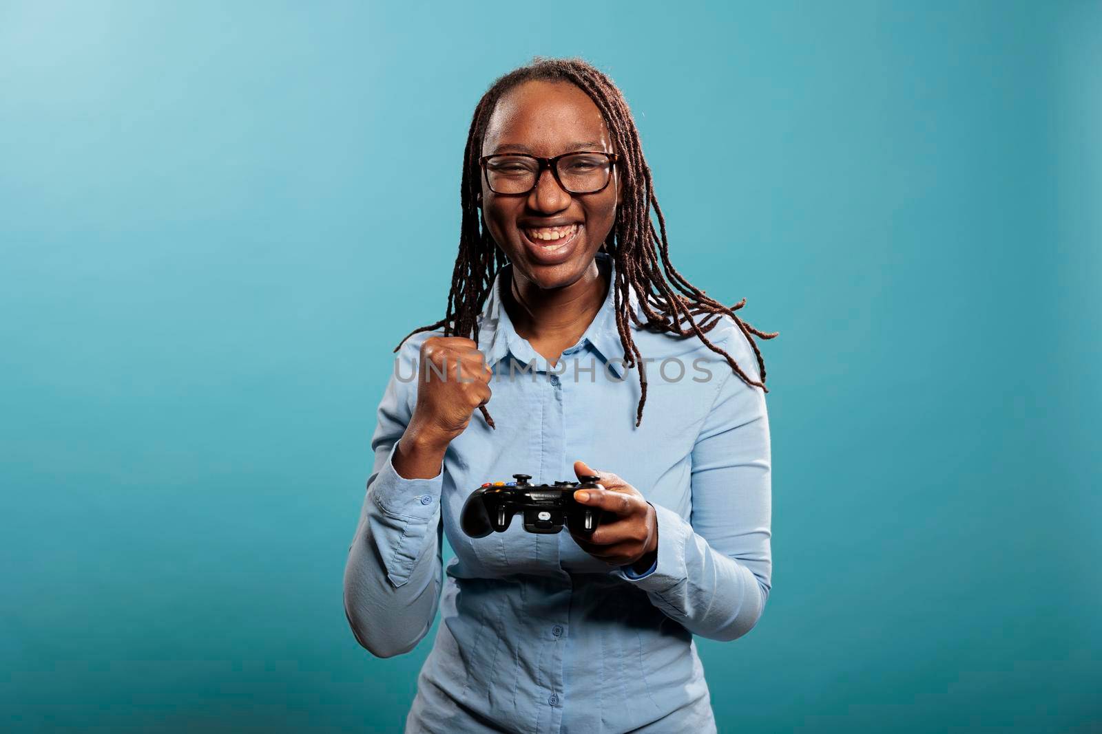 Excited happy woman playing videogames on console while celebrating match victory. Happy joyful young adult person enjoying competitive game win while standing on blue background.