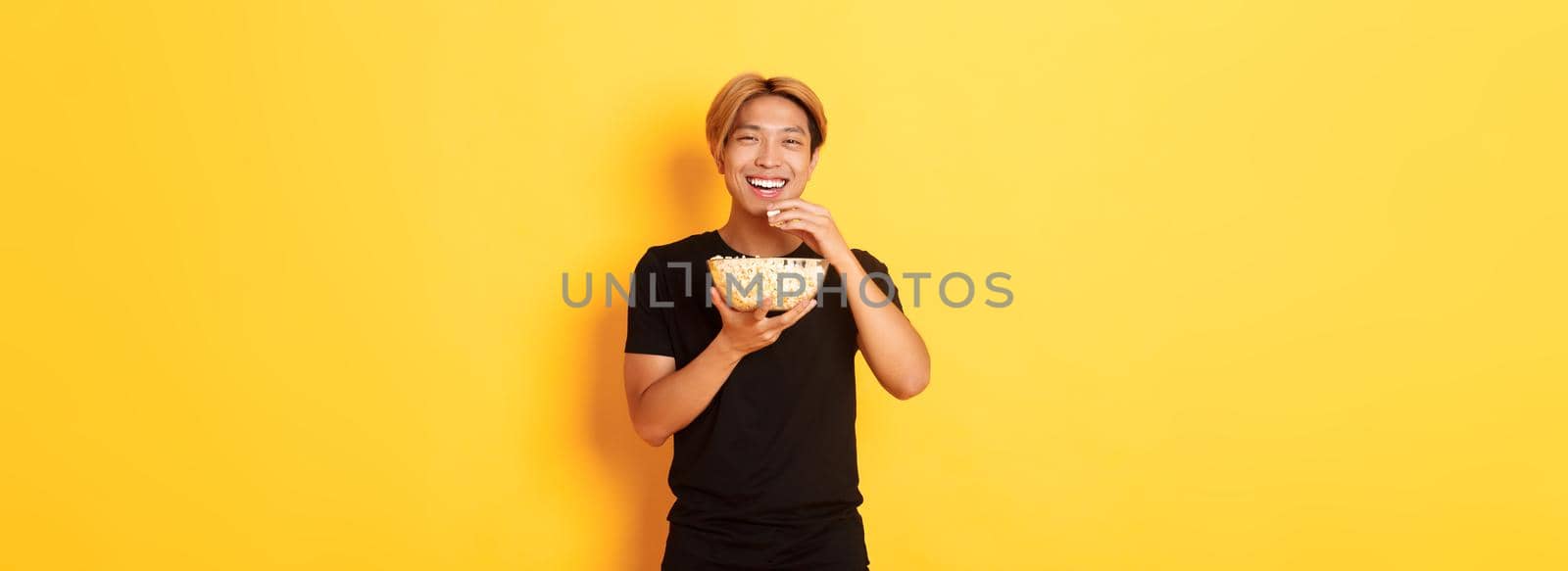 Carefree happy asian guy with blond hair, watching comedy, laughing and eating popcorn, standing yellow background.