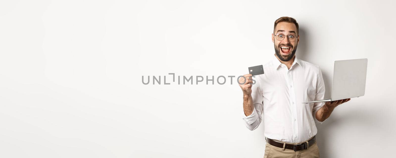 Online shopping. Handsome man showing credit card and using laptop to order in internet, standing over white background by Benzoix