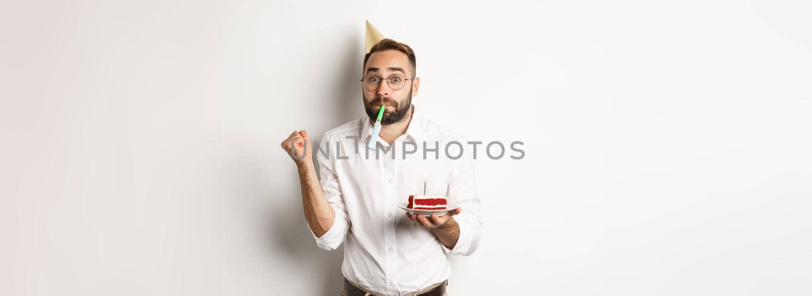 Holidays and celebration. Cheerful man enjoying birthday, blowing party whistle and holding bday cake, white background by Benzoix