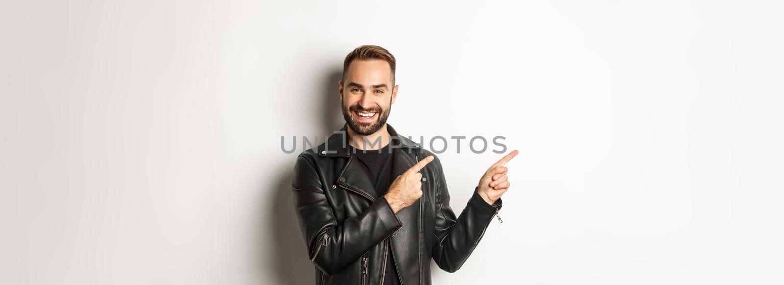 Confident macho man in black leather jacket, pointing fingers left at promo offer, showing logo, white background by Benzoix
