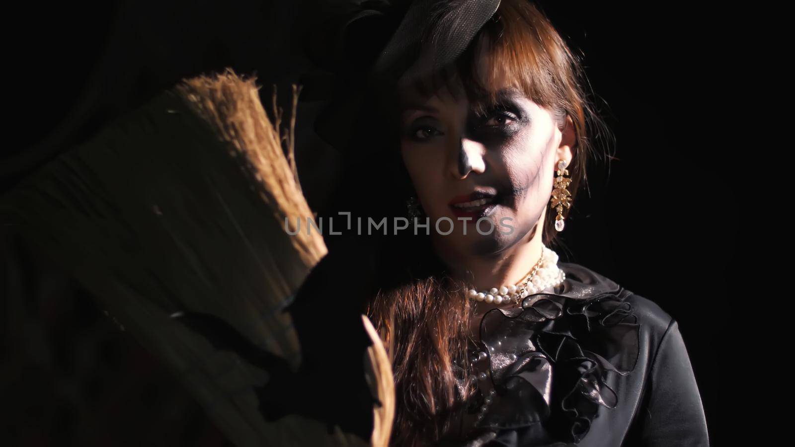 Halloween party, night, frightening portrait of a woman in the twilight, in the rays of light. woman with a terrible make-up in a black witch costume. she holds a broom with a bat by djtreneryay