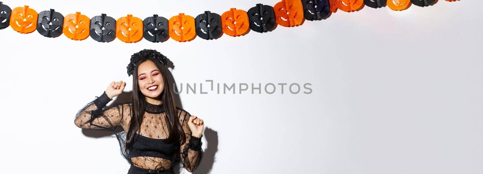 Carefree smiling asian woman in witch costume enjoying halloween party, dancing and rejoicing, standing over white background with pumpkin decoration by Benzoix