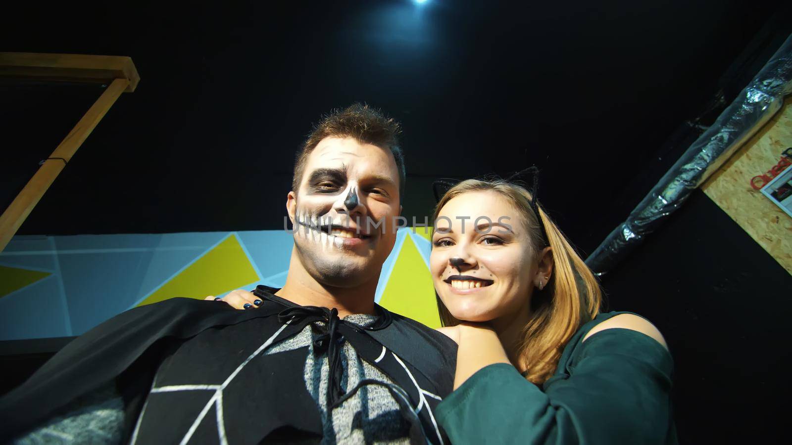 Halloween party, night, frightening portrait of a man with a terrible makeup in a black costume and a girl in a cat costume, they croak in front of the camera. High quality photo