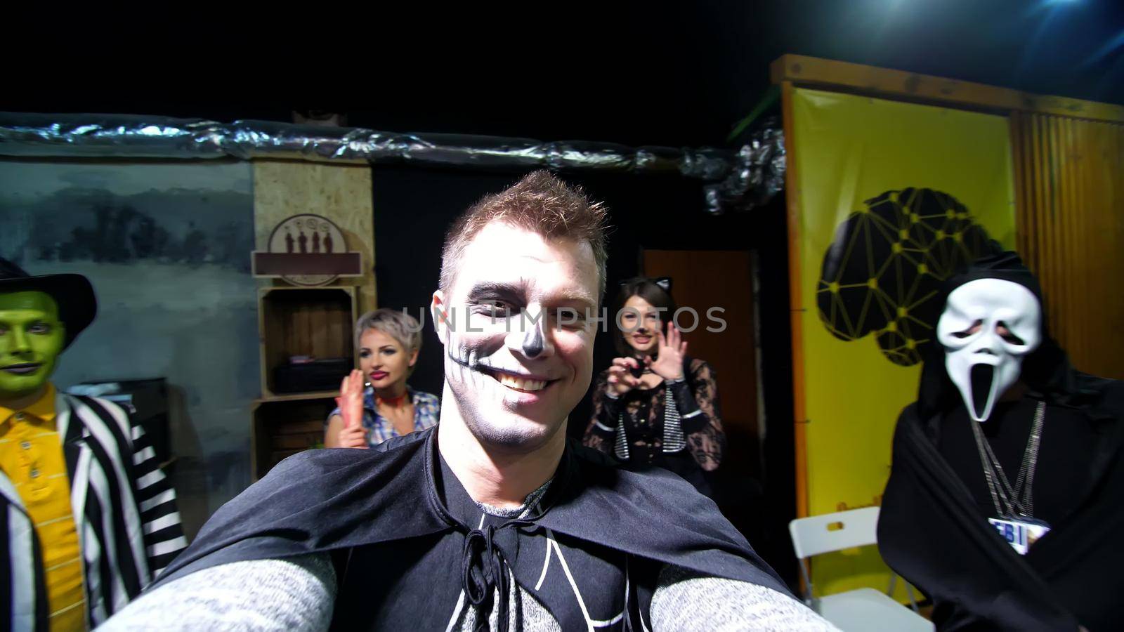 Halloween party, night, frightening portrait of a man with a terrible makeup in a black witch suit, croaks in front of the camera, in the background Halloween scenery is seen. High quality photo