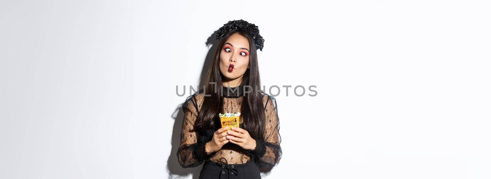 Image of funny carefree girl celebrating halloween in witch costume, making silly faces and holding treats, standing over white background by Benzoix